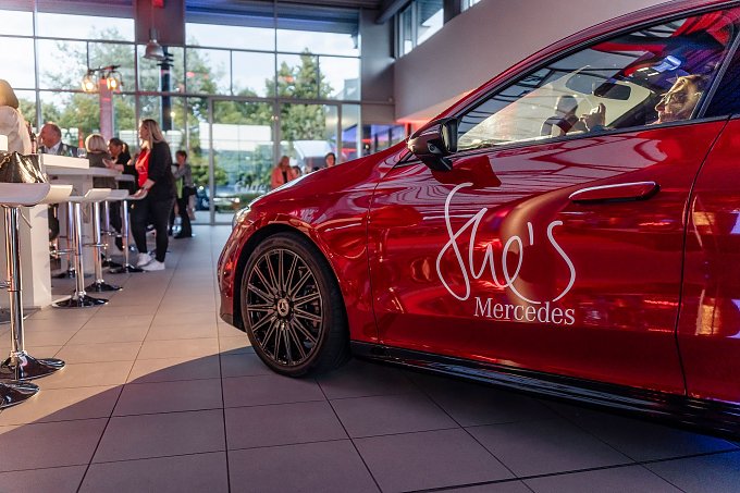Ein rotes Mercedes-Fahrzeug mit der Aufschrift She�s Mercedes auf der Fahrert&uuml;r steht in einem hellen Showroom. Im Hintergrund unterhalten sich Menschen an Stehtischen. Mercedes-Benz CLA 250+ mit EQ-Technologie | Energieverbrauch kombiniert: 14,1-12,2 kWh/100 km | CO2Emissionen kombiniert: 0 g/km | CO2-Emissionsklasse: A